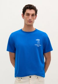 Blå bomulds T-shirt med korte ærmer, der har et hvidt grafisk logo og tekst på venstre bryst. Enkel rund halsudskæring, almindelig pasform.