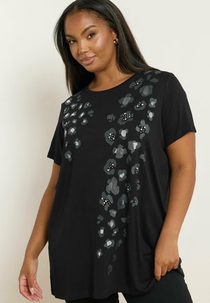 Vrouw in een zwart shirt met korte mouwen, versierd met zilveren en donkergrijze bloemenpatronen en kleine kraalversieringen, staande tegen een effen achtergrond.