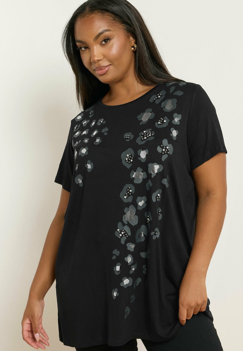 Nainen, jolla on päällään musta lyhythihainen paita, jossa on hopea- ja tummanharmaita kukkakuoseja sekä pieniä helmiä koristeena, seisoo yksinkertaista taustaa vasten.