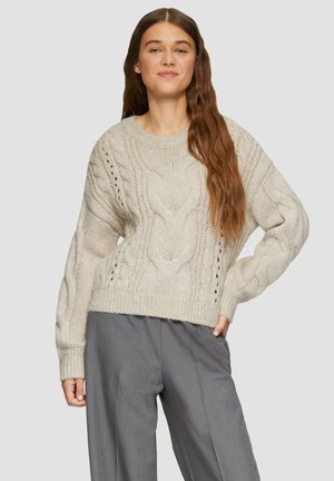 Sweter dziany w jasnym beżu z wzorami w warkocze i otwartymi detalami ściegów. Długie rękawy i ściągacz u dołu. Noszony z szarymi spodniami o szerokich nogawkach.