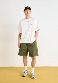 Camiseta blanca de algodón con un pequeño logo negro, pantalones cortos verde oliva, calcetines blancos hasta el tobillo y zapatillas con estampado, con un fondo neutro.