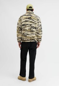 Chaqueta de forro polar con un patrón de rayas de tigre en beige y negro; cuello alto y corte relajado. Combinada con pantalones negros y botas marrones.