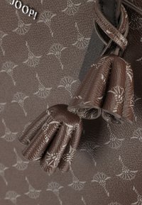Sac à main texturé marron avec un motif floral, orné de pompons et d'un logo argenté. Le design met l'accent sur des matériaux doux et flexibles.