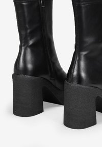 Bottes en cuir noir jusqu'au genou avec un talon texturé massif et une fermeture éclair sur le côté. La finition lisse et les lignes épurées mettent en valeur un design moderne.