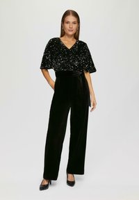 Frau steht mit einer Hand in der Tasche, trägt ein schwarzes, mit Pailletten besetztes Oberteil mit weiten Ärmeln und hochtaillierte schwarze Samenhosen mit Gürtel und Absatzschuhen.