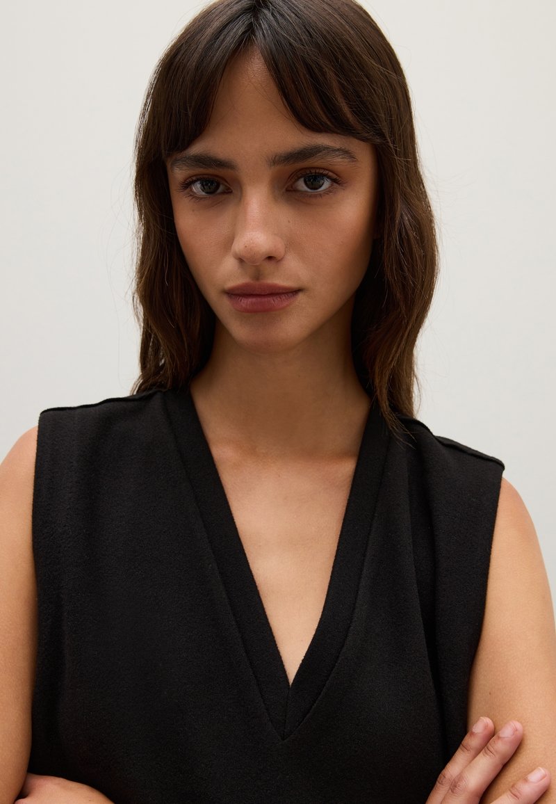 Ärmelloses schwarzes Kleidungsstück mit V-Ausschnitt, aus strukturiertem Stoff gefertigt. Das Design zeichnet sich durch eine lockere Passform mit dezenten Schulterakzenten aus.