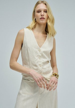 Mujer rubia que lleva un chaleco beige sin mangas abotonado y pantalones a juego, y accesorios con pendientes de aro dorados y una pulsera dorada gruesa.