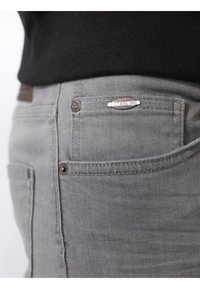 Jeans de mezclilla gris con una textura suave, que presentan una placa de logo visible y bolsillos traseros estándar. Vista en primer plano del área de la cintura.