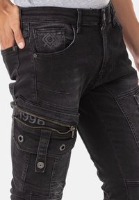 Des pantalons cargo en denim noir présentent une coupe ajustée, des poches zippées et des coutures décoratives avec un détail de 1996 près de la fermeture éclair.