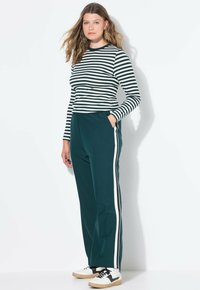 Jongeman met lang haar, gekleed in een zwart-wit gestreepte lange mouwen top, donkere trainingsbroek met witte zijstrepen en witte sneakers.