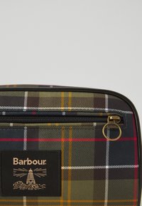 Tartánmintás Barbour táska sarka fekete cipzárral és sárgaréz cipzárhúzóval, fekete folttal, amely világítótorony logót és a márkanevet tartalmazza.
