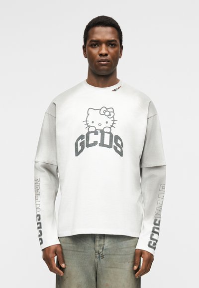 Άνδρας που φοράει λευκό φούτερ με μαύρο γραφικό Hello Kitty και κείμενο «GCDS», στέκεται μπροστά σε ένα απλό ανοιχτό φόντο.