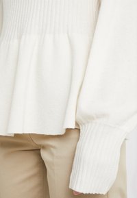 Bílý svetr s peplum designem a žebrovanými manžetami, kombinovaný s béžovými kalhotami, vytváří jemný a neutrální outfit.