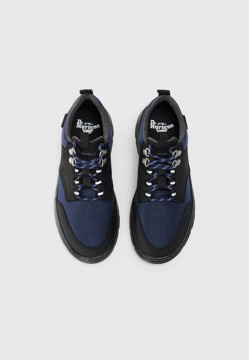 Et par navy blue og sorte Dr. Martens-sko med reflekterende snørebånd, metaløjner og polstret indre foret, vist fra oven på hvid baggrund.