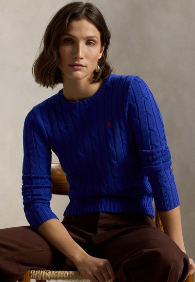 Blå kabelstrikket genser med ribbestrikket halslinning og mansjetter. Har en liten rød logo på brystet. Kombinert med brune bukser.