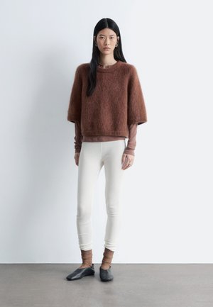 Bruin, pluizig gebreid cropped truitje over een aansluitende lange-mouwen top, gecombineerd met witte, gestructureerde leggings en zwarte platte schoenen. Eenvoudig, modern ontwerp.