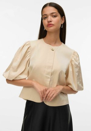 Žena v béžovej blúzke s nafúknutými rukávmi a zlatým gombíkom, v čiernej sukni, stojaca so zloženými rukami na bielom pozadí.