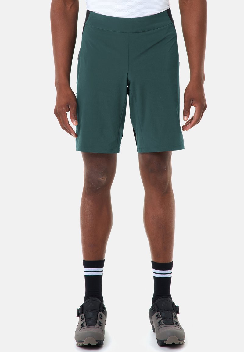 Homme portant un short de sport vert foncé, des chaussettes rayées noires et blanches, et des chaussures de cyclisme grises, debout devant un fond blanc.