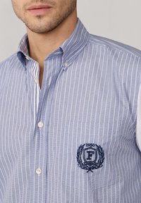 Uomo che indossa una camicia a righe verticali blu e bianca con un colletto a bottoni e un ricamo con la scritta "1950 F" sulla tasca del petto.