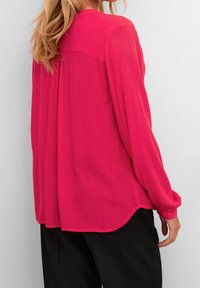 Blouse à manches longues en tissu rose vif, présentant une coupe décontractée, un détail plissé au dos et un ourlet arrondi, associée à un pantalon noir.