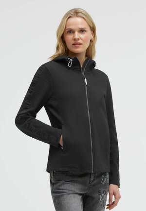 Woman wearing a black textured zip-up hoodie with text on sleeves and gray jeans with printed design on front thigh.