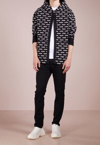 Man i svart zip-up huvtröja med vit "VERSUS" tryck, vit t-shirt, svarta byxor och vita sneakers som står på ett beige golv.