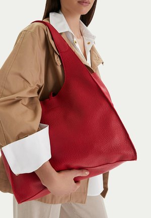 Mujer con chaqueta beige y camisa blanca sostiene un gran bolso rojo de cuero texturizado sobre el hombro, con fondo liso.