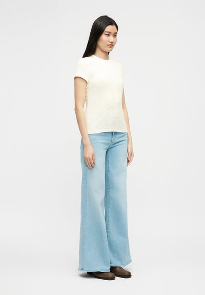 Γυναίκα που στέκεται φορώντας μια κοντομάνικη κρεμ κορυφή, ανοιχτόχρωμο μπλε τζιν με φαρδιά πόδια και καφέ παπούτσια, μπροστά σε απλό ανοιχτό φόντο.