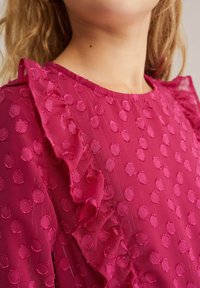 Ženska v roza teksturirani bluzici z dvignjenimi pikami in nabranimi detajli okoli ramena in ovratnika.