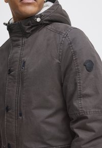 Chaqueta con capucha gris de tela texturada, con forro de piel sintética, botones a presión, un bolsillo en el pecho con cremallera y un parche con el logo en la manga.