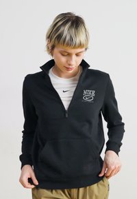 Melna Nike sporta jaka ar ceturtdaļas rāvējslēdzēju, priekšējo kabatu un logo uz krūtīm. Gluda tekstūra, garas piedurknes un nedaudz pieguļošs dizains.