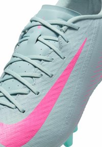 Fotbalová kopačka Nike v světle modré barvě s texturovaným povrchem, vybavená jasně růžovým logem Swoosh, plochými tkaničkami a kolíky v akvamarínové barvě.