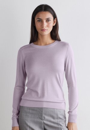 Frau mit langen dunklen Haaren, die einen hellvioletten Langarmpullover und graue Hosen trägt und vor einer schlichten Wand steht.