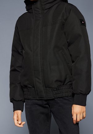 Person iført en sort polstret jakke med elastiske manchetter, forlommer og Tommy Hilfiger-logo på brystet og ærmet.