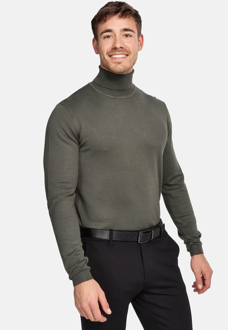 Olivgrüner Rollkragenpullover aus Rippstrick, kombiniert mit schwarzen Hosen und einem schwarzen Gürtel. Enganliegende Silhouette, lange Ärmel.