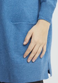 Niebieski dziany sweter z prążkowanym ściągaczem u dołu i przy mankietach, bocznymi rozcięciami oraz przednią kieszenią. Gładka faktura o średniej grubości.