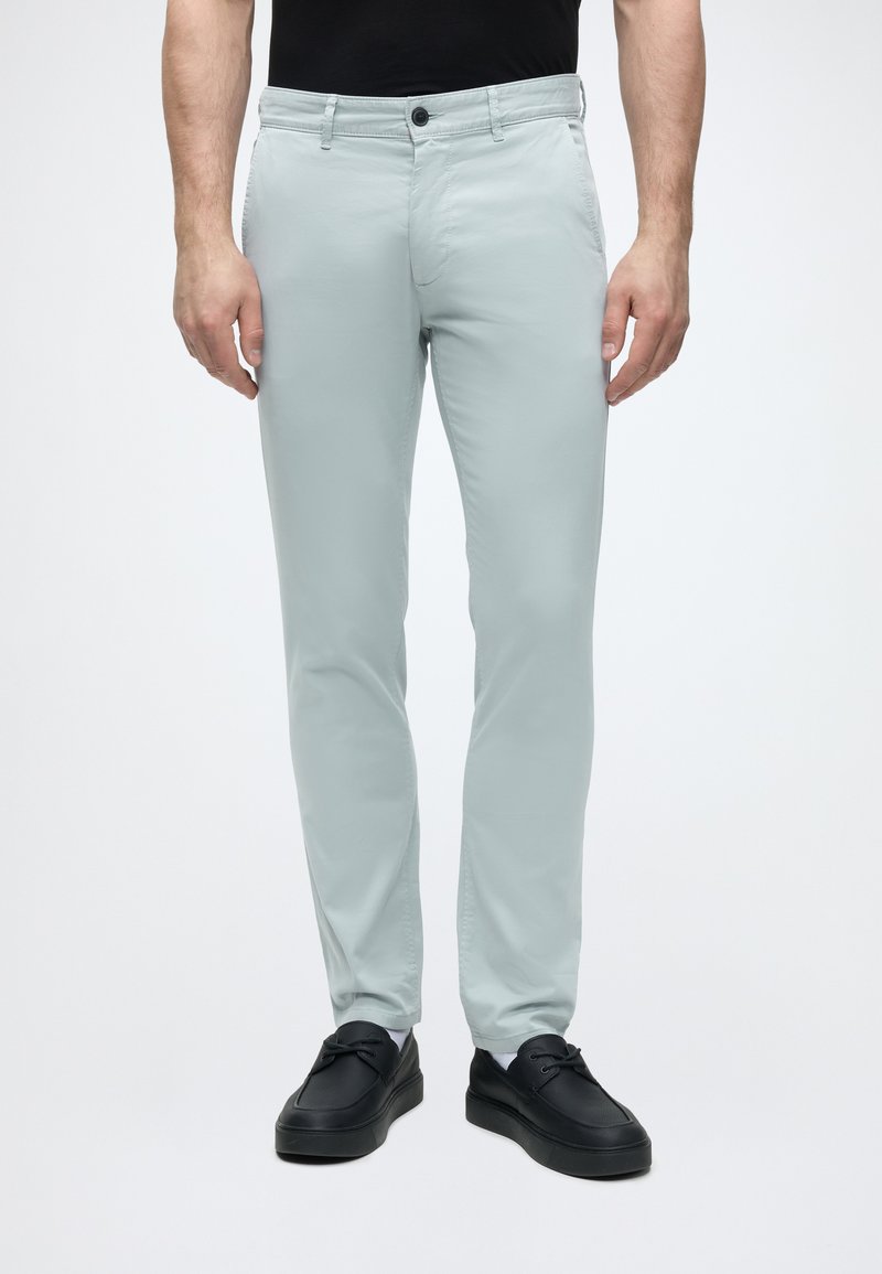 Homme portant un pantalon slim gris clair et des chaussures en cuir noires, debout devant un fond blanc uni.
