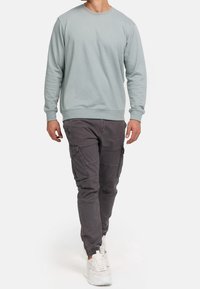 Hombre vestido con sudadera gris clara, pantalones cargo gris oscuro con bolsillos y zapatillas blancas caminando frente a un fondo blanco.