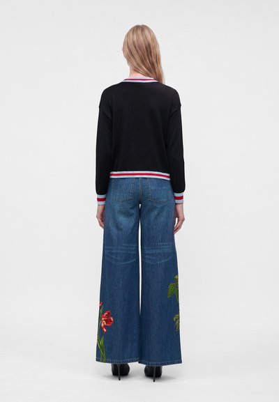 Femme aux longs cheveux blonds, de dos, portant un pull noir avec des poignets rayés et un jean bleu large avec une broderie florale.