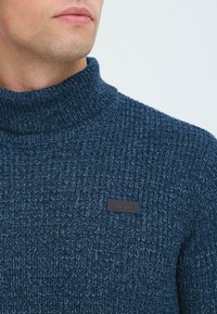 Pleteni navy pulover s turtleneck ovratnikom in teksturiranim vzorcem, na prsih pa je majhna črna usnjena oznaka. Oblačilo s kragnim oblikovanjem in povišanim ovratnikom.