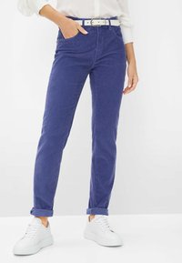 Pantaloni di velluto a coste viola con una vestibilità slim, orli risvoltati e tasche; abbinati a una cintura bianca e sneaker bianche.