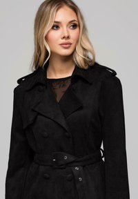 Czarny zamszowy prochowiec z dwurzędowym zapięciem, dużymi guzikami, paskiem w talii i szerokimi klapami.