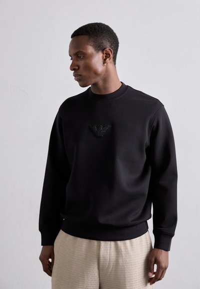 Schwarzer Sweatshirt mit Rundhalsausschnitt, ausgestattet mit einem strukturierten schwarzen Logo-Patch auf der Brust. Glattes Material und lässige Passform.