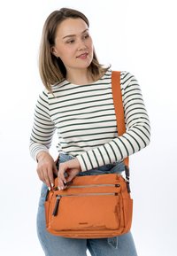 Young woman wearing a white and green striped top and blue jeans, adjusting the zipper of an orange crossbody bag.