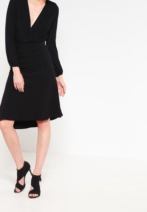 Kobieta ubrana w czarny top z dekoltem w serek i długim rękawem, czarną spódnicę typu high-low oraz czarne, otwarte buty na wysokim obcasie, stojąca na białej podłodze.