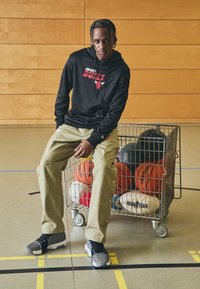 Sweatshirt à capuche noire avec impression rouge, pantalon cargo beige avec boutons-pression, et chaussures de sport grises, assis à côté d'un chariot rempli de divers ballons de basket.