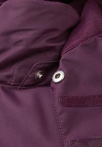 Tessuto giacca bordeaux con texture liscia, dotata di un bottone a scatto in metallo, dettaglio in velcro e rivestimento in pile di contrasto sul colletto.