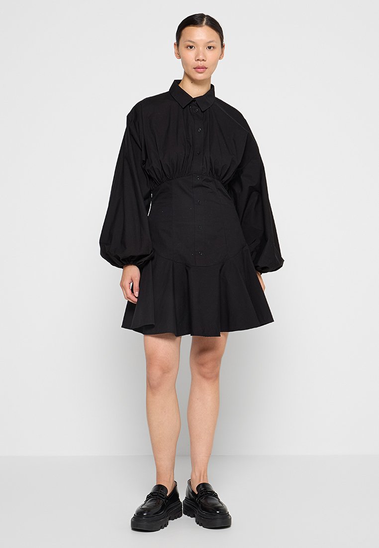 Frau in einem schwarzen kurzärmligen Kleid mit langen Puffärmeln, tailliertem Schnitt, ausgestelltem Rock und schwarzen klobigen Slippern, die vor einem schlichten Hintergrund steht.