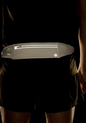 Reflekterende gråt løbebælte med lynlås, båret rundt om taljen over sorte atletiske shorts og skjorte på en person.