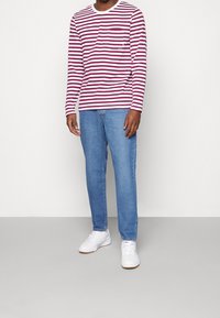 Mann in einem rot-weißen gestreiften Langarmshirt, blauen Jeans und weißen Turnschuhen stehend vor einem einfarbigen Hintergrund.